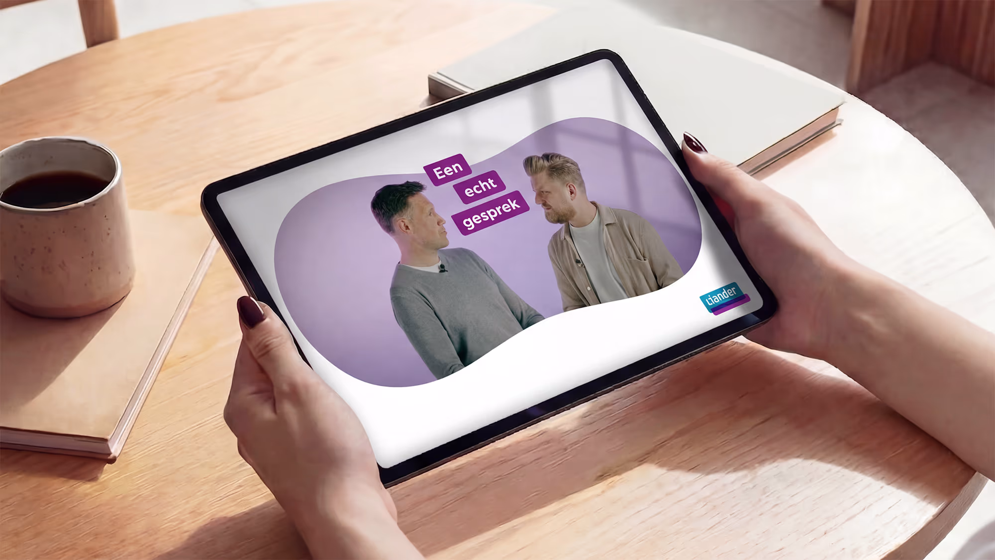 Tablet screen showing two men in conversation with the text 'Een echt gesprek' and a Liander logo on a wooden table beside a coffee cup and a notebook.
