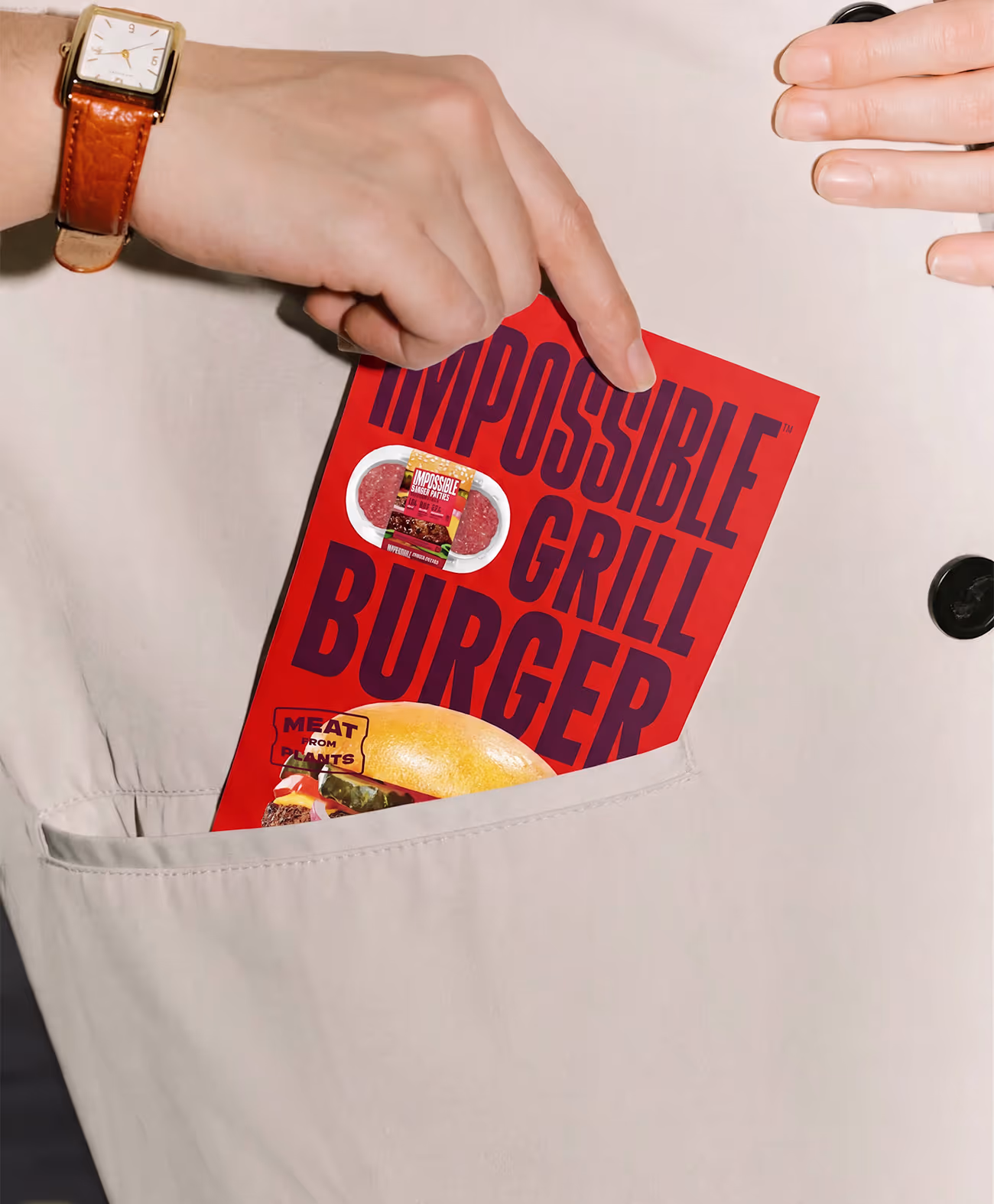 Hand placing a red Impossible Grill Burger package with plant-based meat into a beige coat pocket.