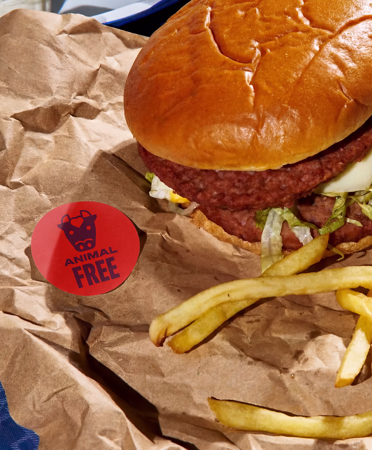 Plant-based burger with lettuce and tomato in a bun, accompanied by French fries on brown paper with a red sticker labeled 'Animal Free'.
