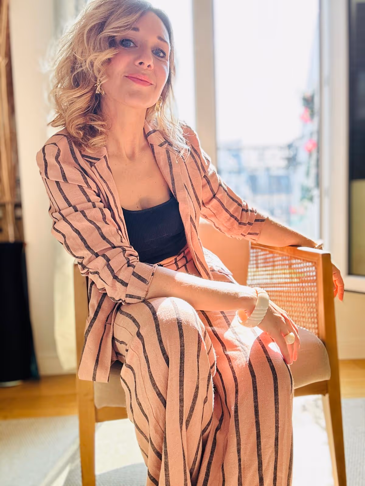 Une femme aux cheveux blonds bouclés, assise sur une chaise en bois, vêtue d'un tailleur à rayures roses et d'un haut noir.