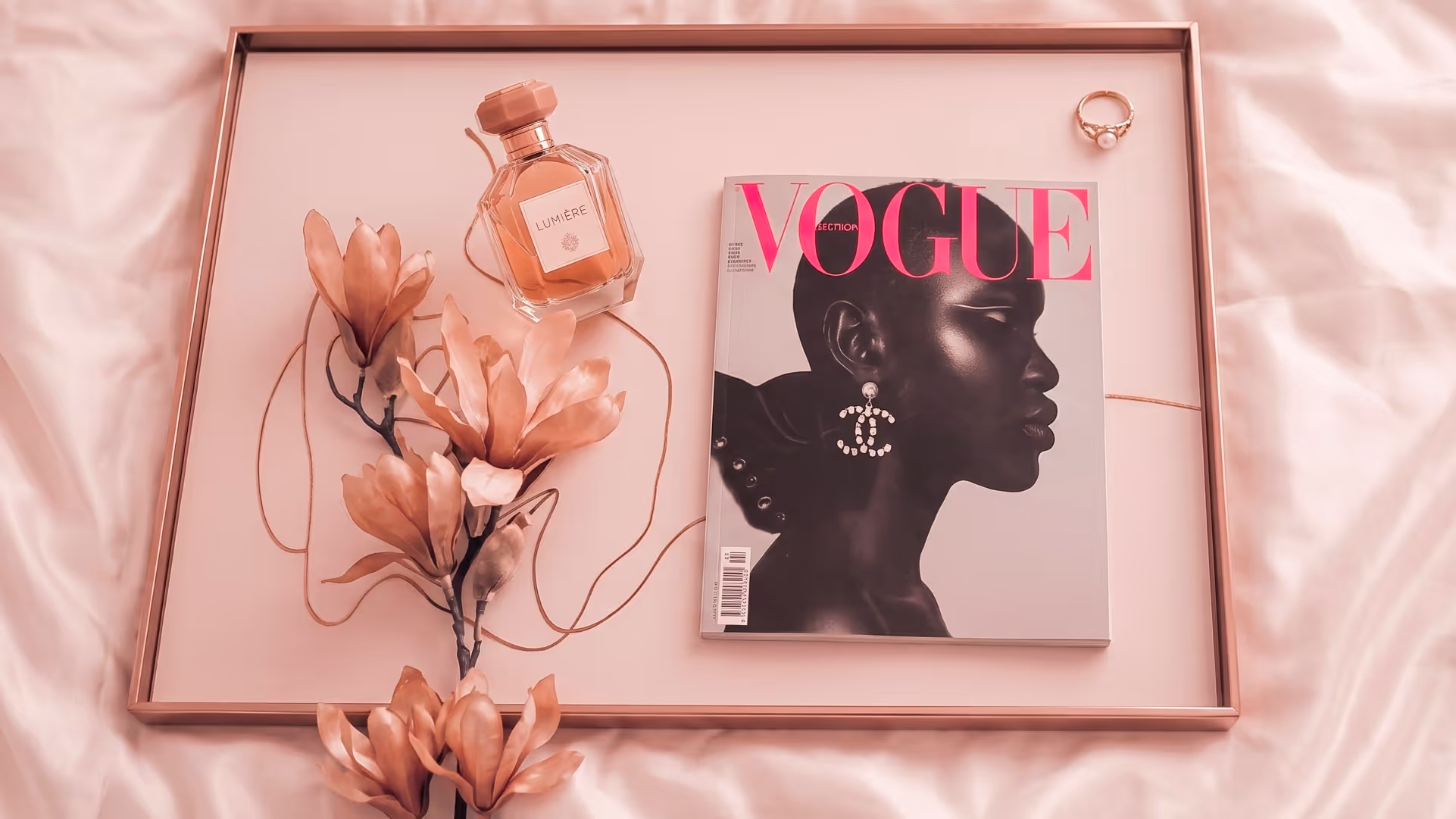 Vogue magazine with profile of woman, perfume bottle, pink flowers, and pearl ring on satin tray.