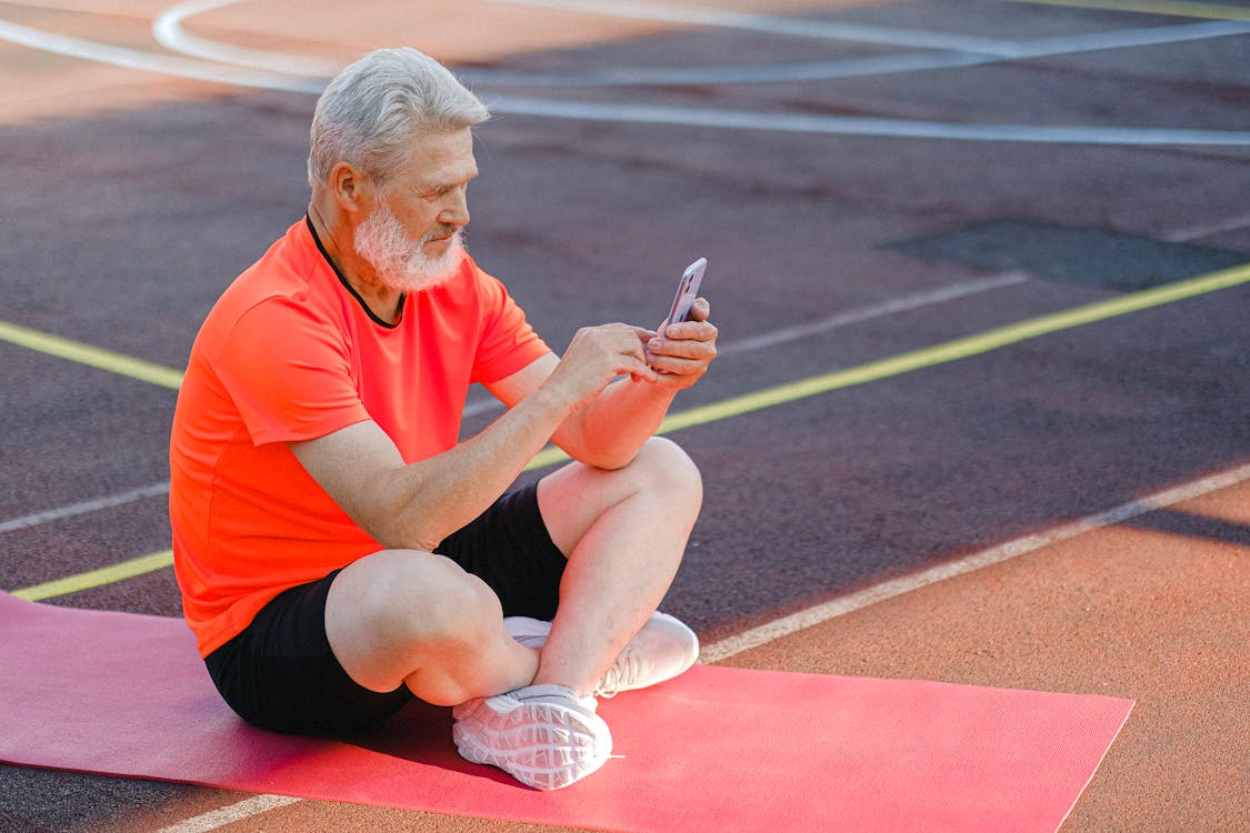  Full body of focused senior male athlete in sportswear sitting on stadium and browsing mobile phone during training Stock Photo