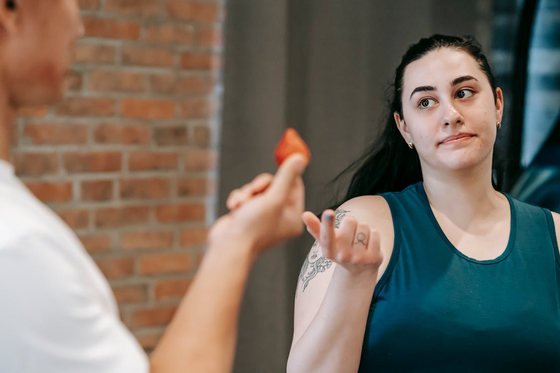 Free Unrecognizable personal coach offering ripe berry to ethnic overweight female in activewear while helping to lose weight in gym near brick wall Stock Photo