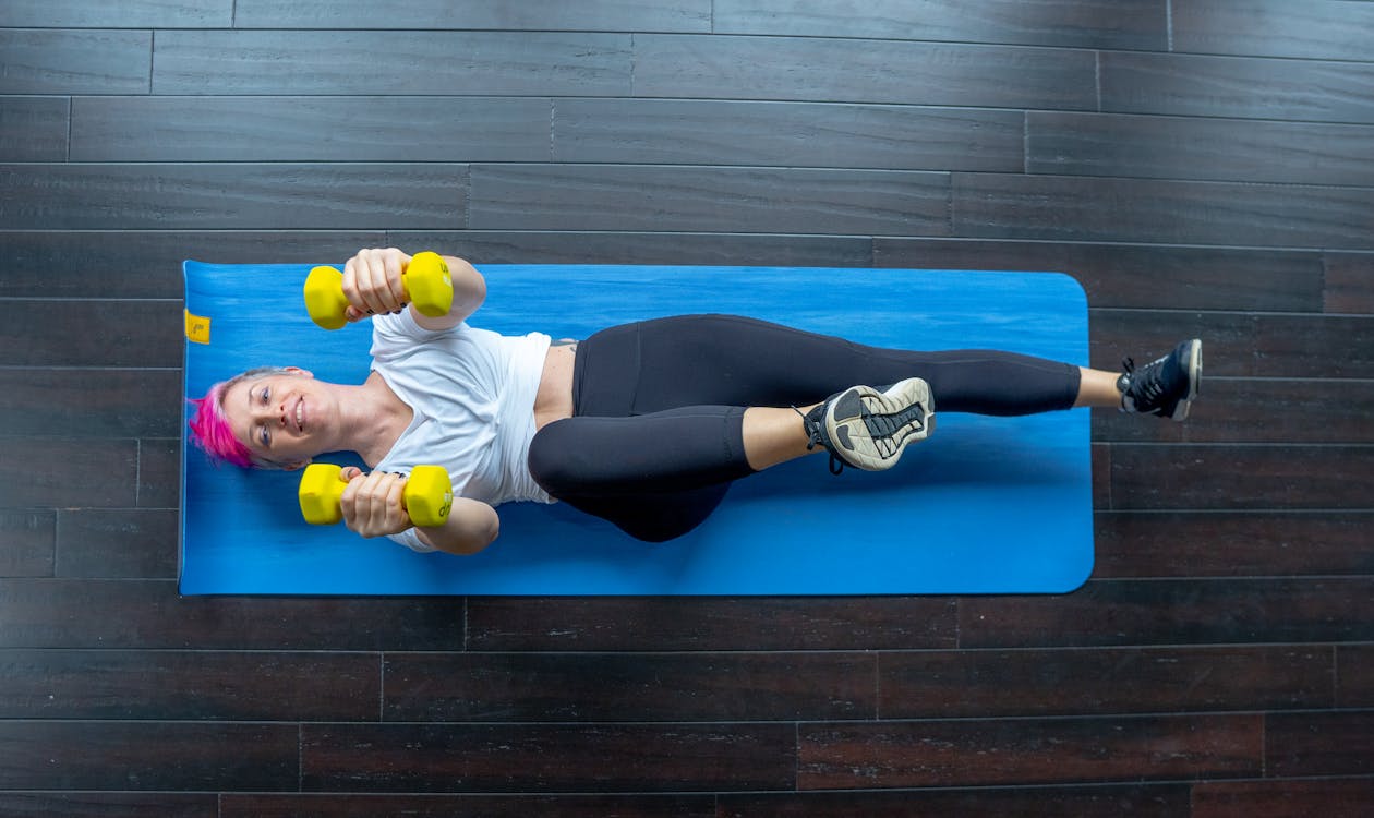 Free Woman in White T-shirt Exercising While Lying Down on a Yoga Mat Stock Photo