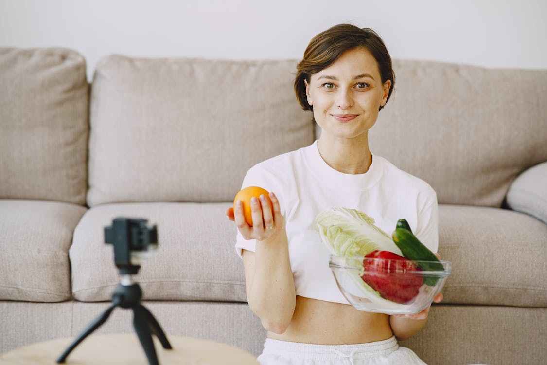 Free Optimistic female in white clothes sitting near sofa and holding bowl with fresh vegetables and orange in hands and looking at camera Stock Photo