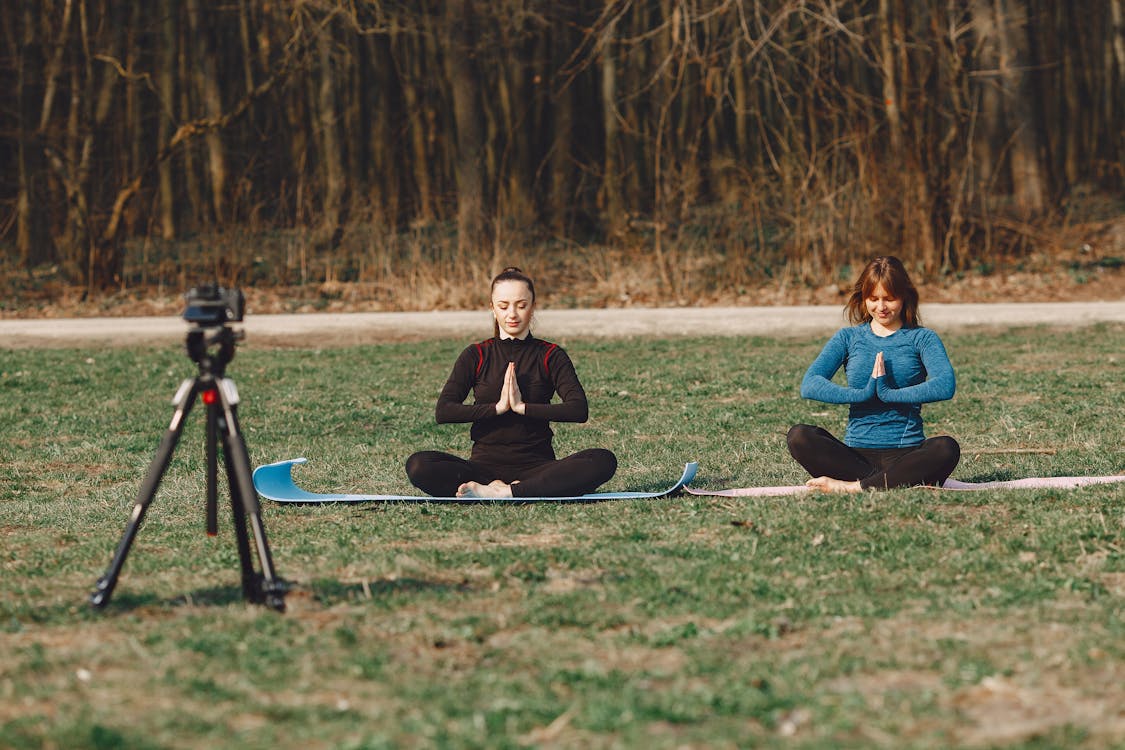 Free Calm young female friends in sportswear sitting with crossed legs and closed eyes in Namaste pose in front of tripod with photo camera on grass Stock Photo