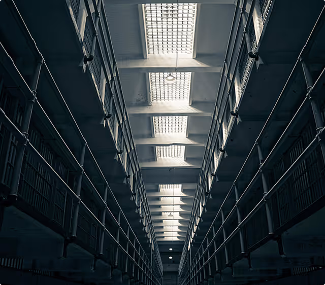 Interior view of a dimly lit prison corridor with metal railings and barred cells on both sides.