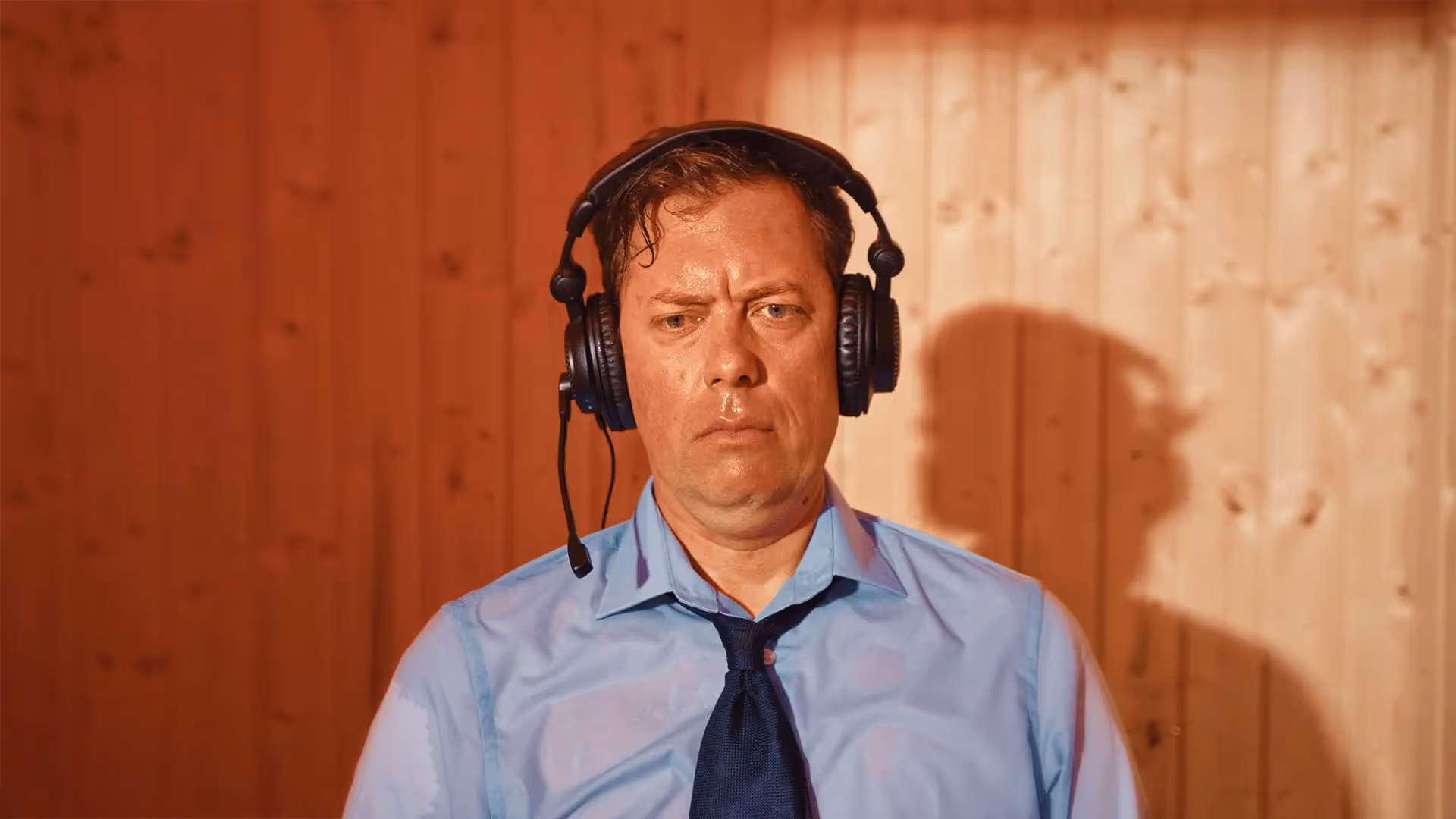 Man met een blauw overhemd, das en zwarte headset met microfoon, kijkt serieus met houten wand op de achtergrond.