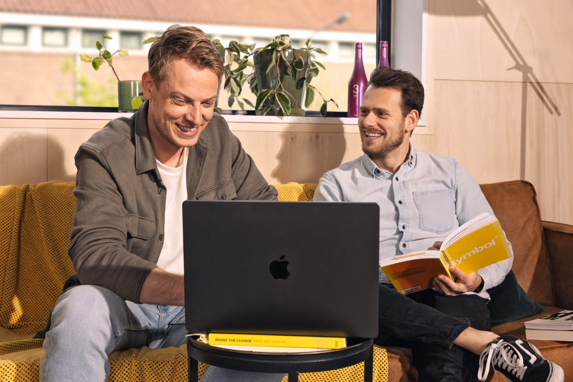 Twee mannen zitten op een bank, de een werkt op een laptop en de ander leest een boek met de titel 'Symbol'.