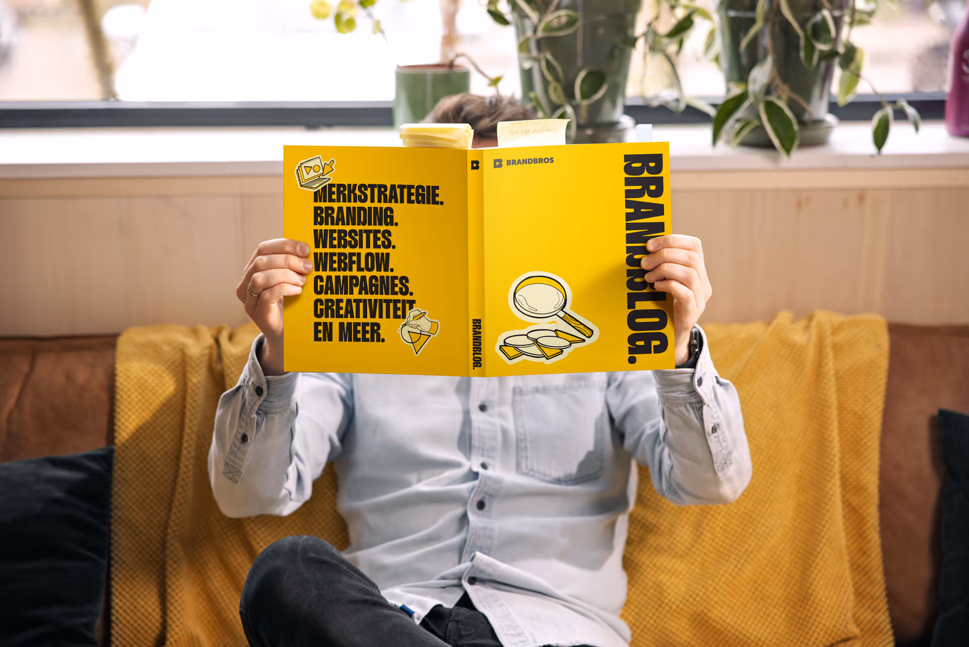 Persoon zittend op een bank leest een felgeel boek met de titel 'BRANDBLOG' en tekst over merkstrategie en branding.