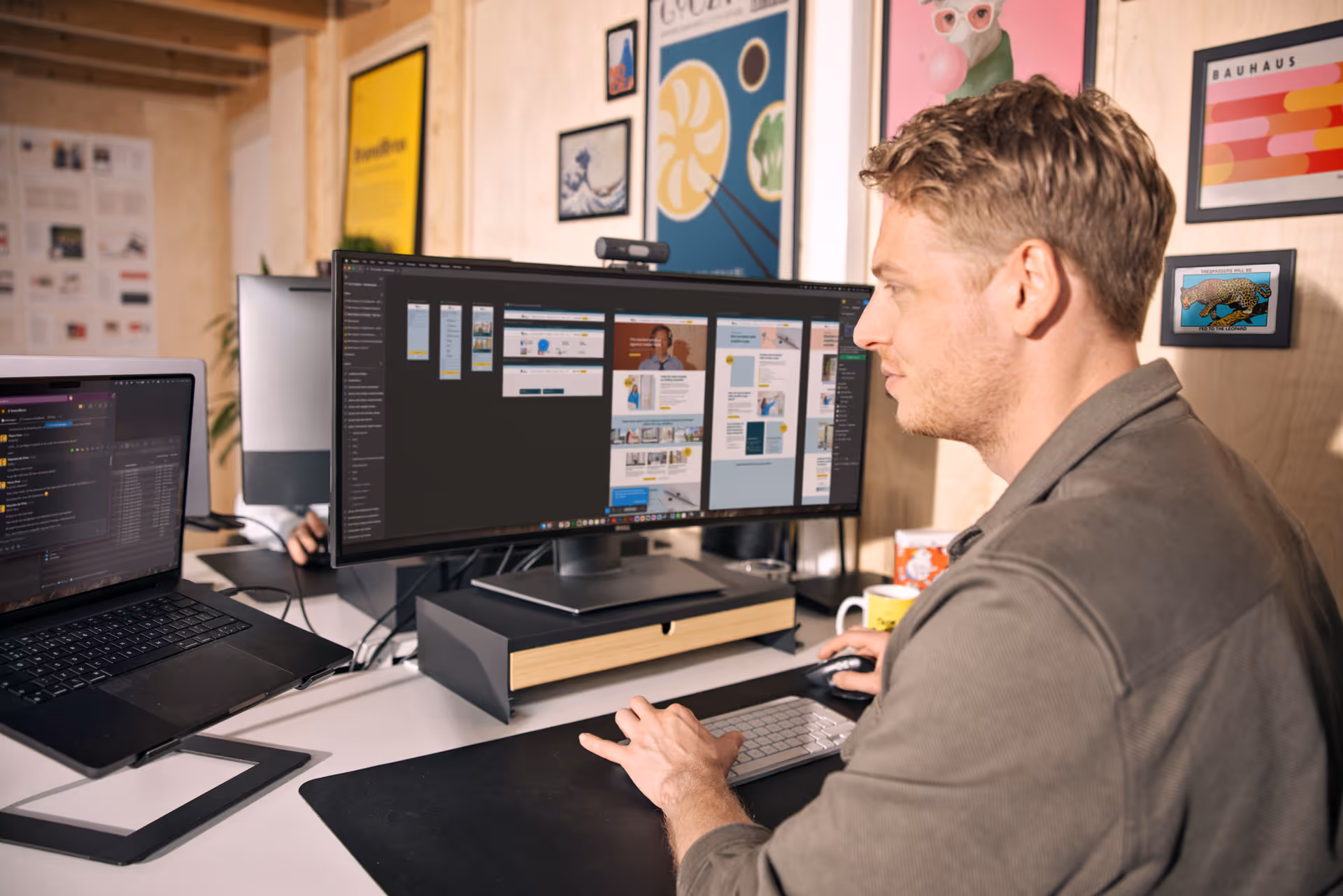 Man werkt aan een bureau met twee beeldschermen, een laptop en een grote monitor met ontwerpsoftware, omringd door kleurrijke posters.