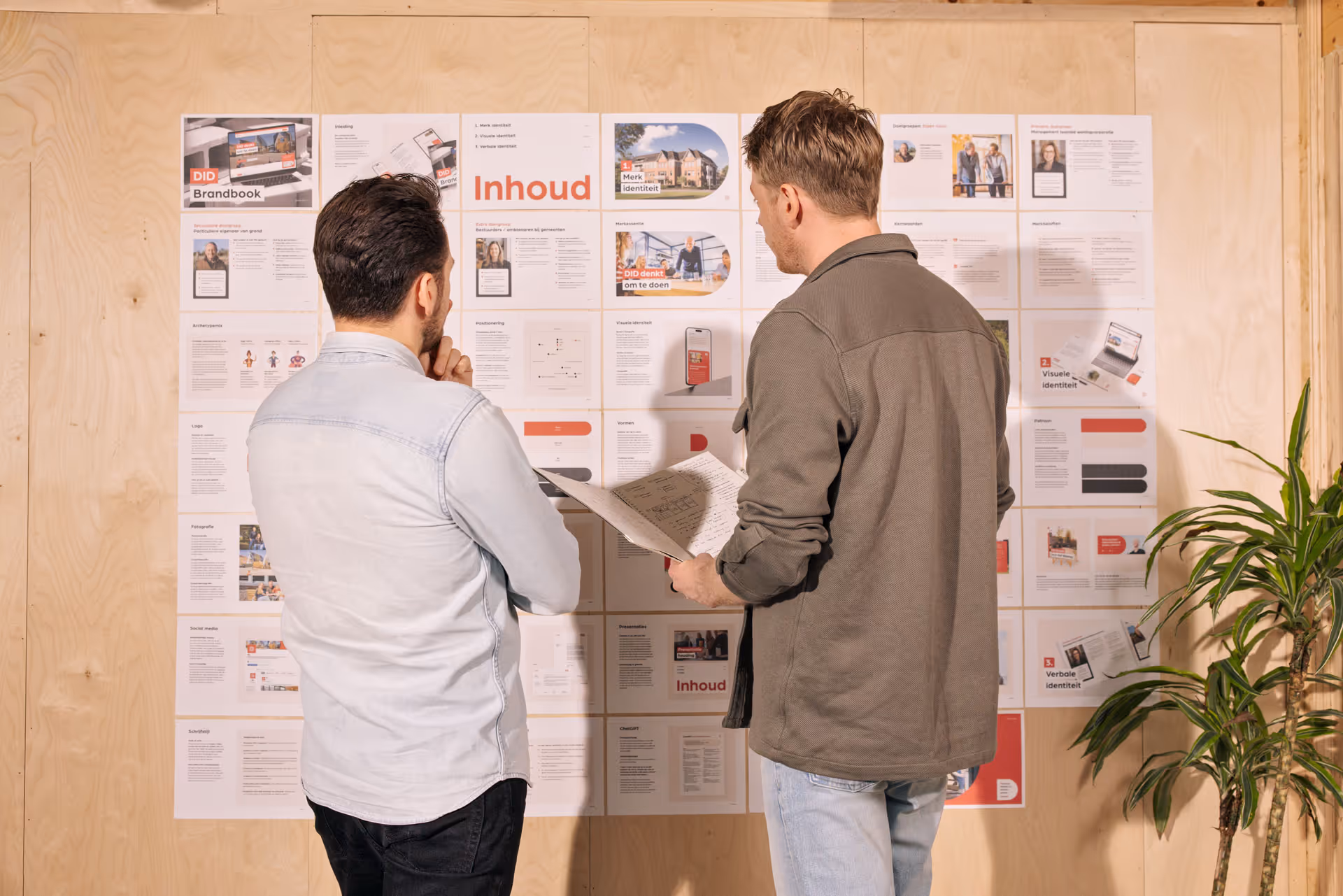 Twee mannen bespreken documenten terwijl ze naar een muur vol met een brandbook en inhoudsopgave kijken.