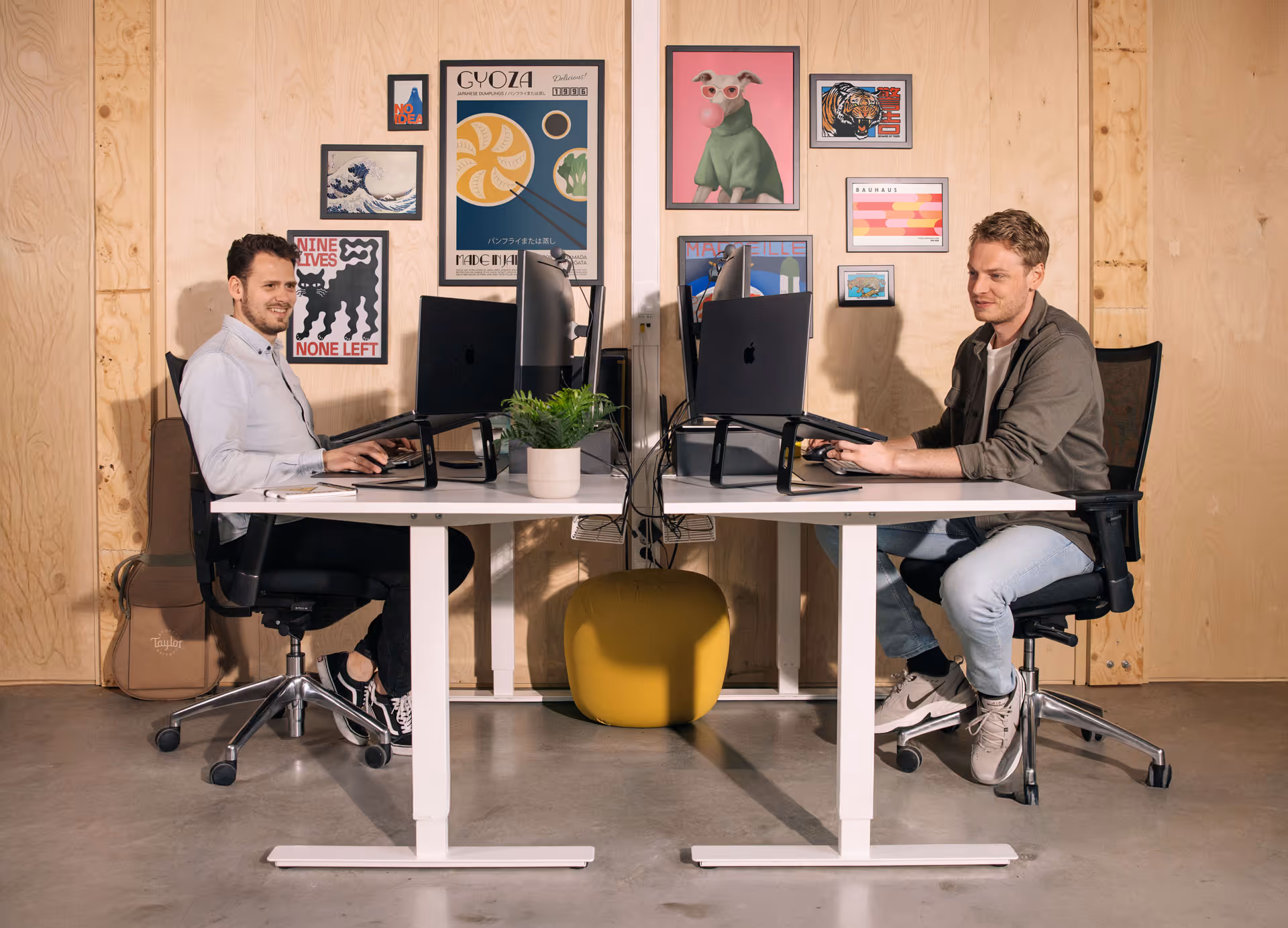 Twee jonge mannen zitten aan tegenovergestelde zijden van een gedeeld bureau met computers in een moderne kantoorruimte met houten wanden en kleurrijke posters.