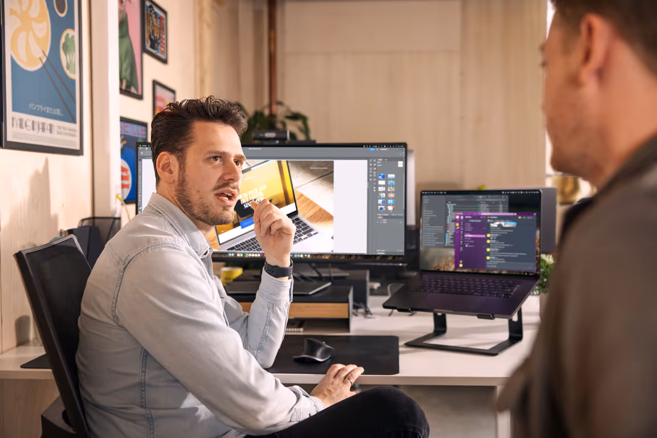 Man in een casual overhemd bespreekt iets zittend aan een bureau met twee beeldschermen en een laptop in een kantooromgeving.