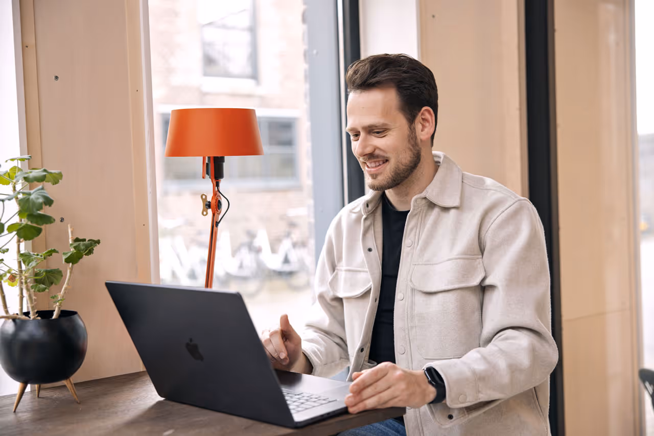 Man met beige jas zit aan houten tafel en lacht tijdens videogesprek op laptop bij raam.