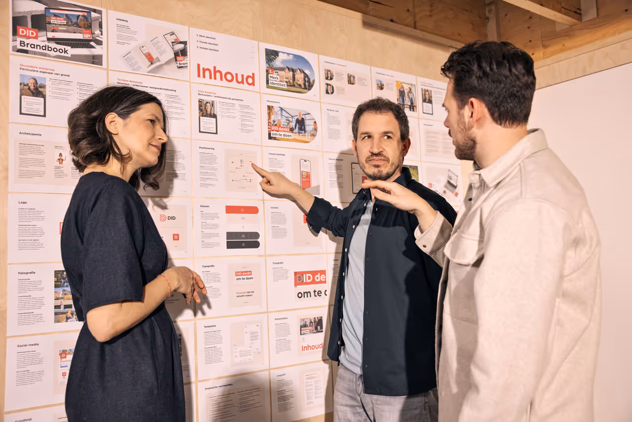 Drie personen bespreken en wijzen naar een wand met uitgestalde did-brandboekpresentaties en inhoudsopgave.