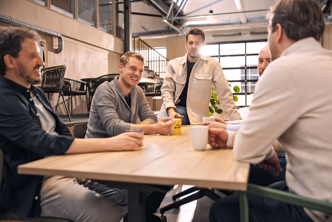 Vijf mannen die bij een houten tafel in een industrieel kantoor zitten en koffie drinken tijdens een informele bespreking.