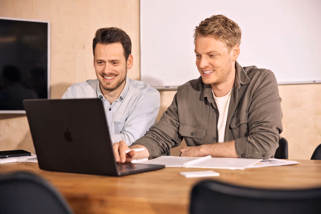Twee mannen die samen lachend op een laptop kijken aan een houten tafel met papieren en notitieboekjes.