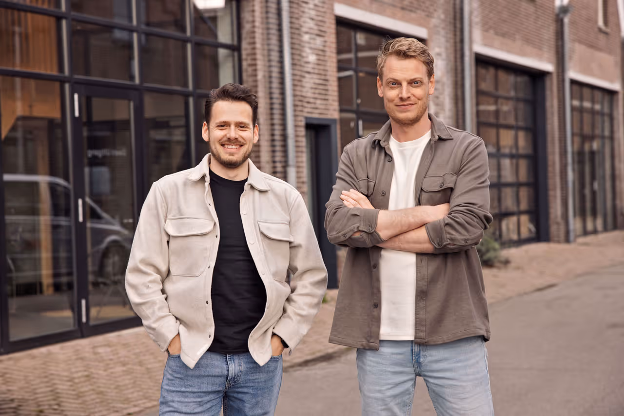 Twee mannen staan lachend voor een gebouw met grote ramen en bakstenen gevel.
