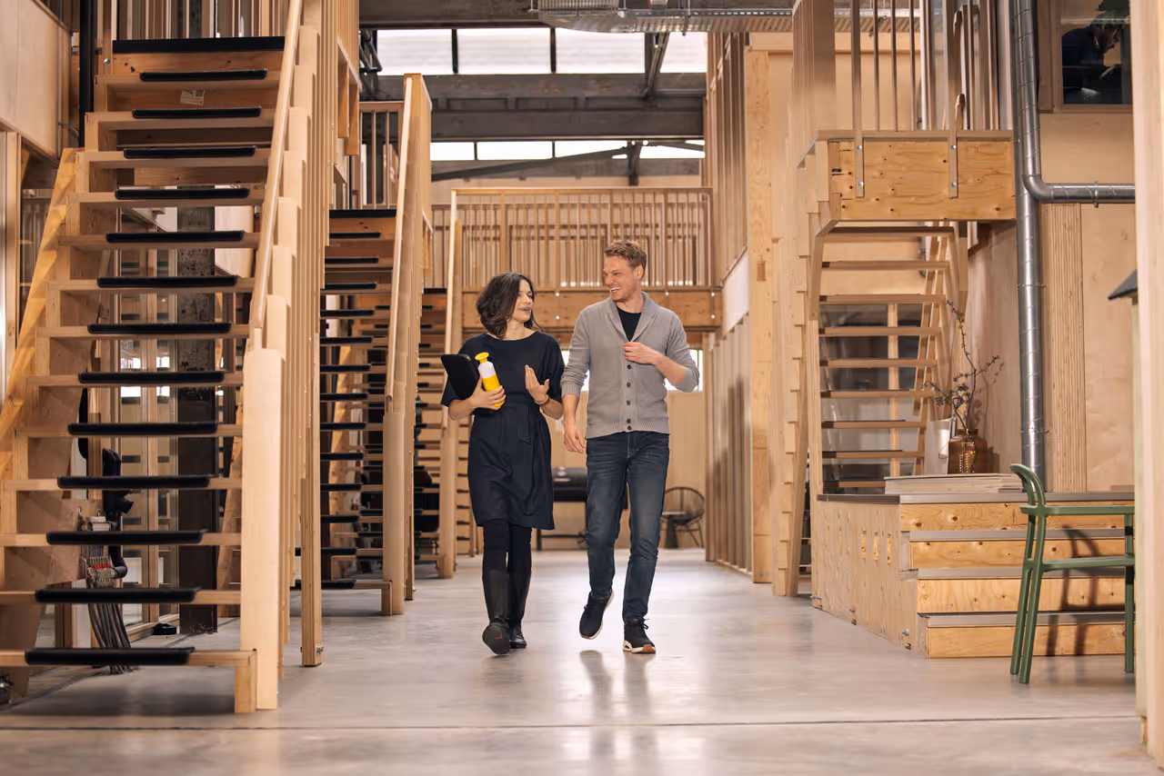 Twee mensen lopen en praten door een ruime, lichte gang met houten trappen en moderne inrichting.