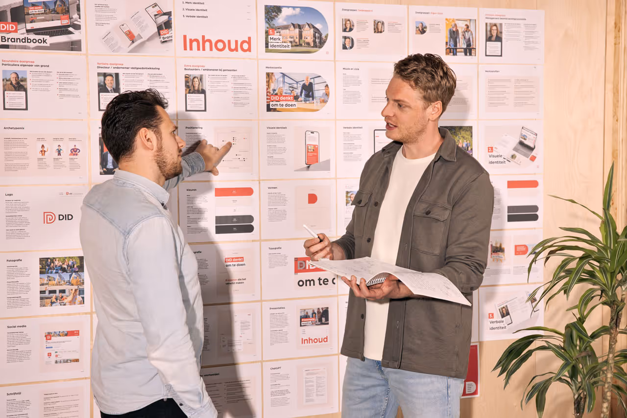 Twee mannen bespreken en wijzen naar uitgeprinte presentatiepagina's over branding op een muur.