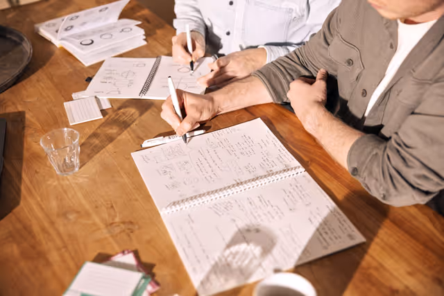 Twee personen schrijven in schetsboeken en notities met diagrammen en aantekeningen aan een houten tafel.
