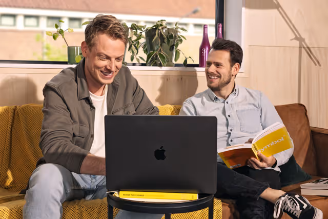 Twee mannen zitten op een gele bank, een van hen werkt op een MacBook terwijl de ander met een boek 'symbol' in zijn hand lacht.