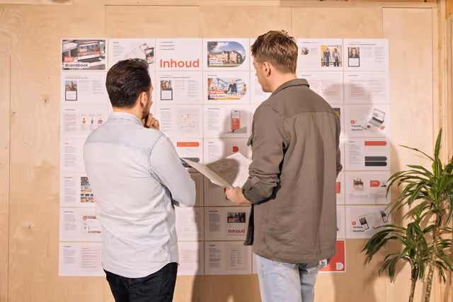 Twee mannen bekijken en bespreken een muur met geplaatste documenten en afbeeldingen voor een brainstormsessie.