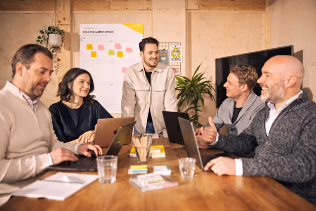 Vijf collega’s bespreken ideeën in een moderne vergaderruimte met laptops en notities op tafel.