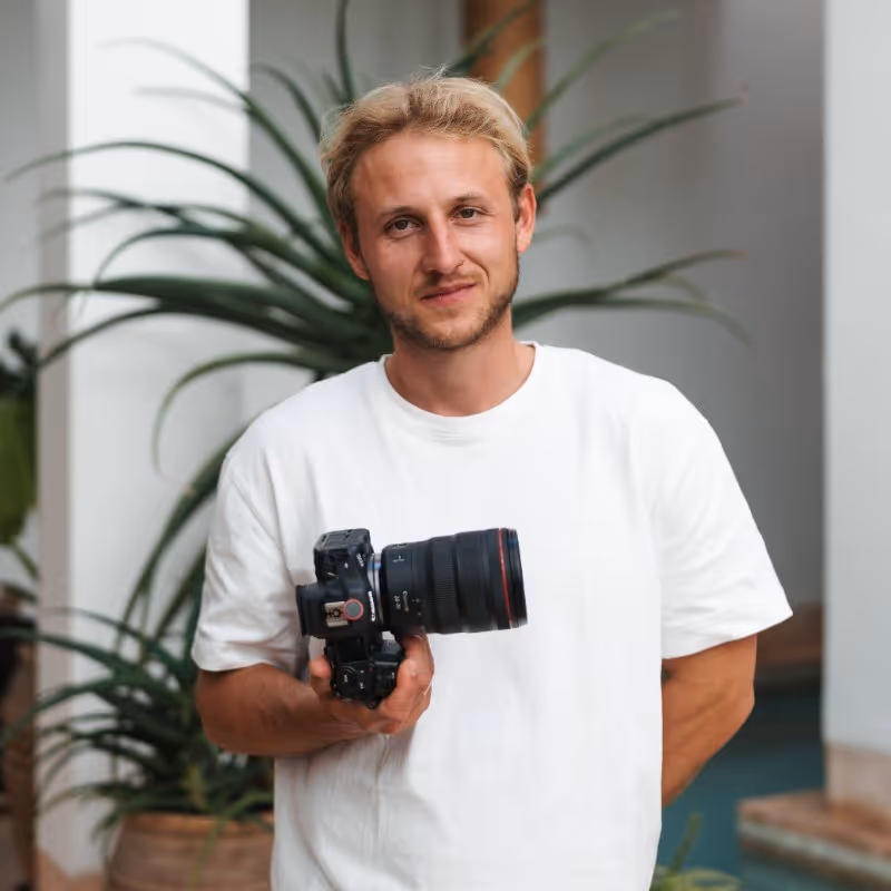 Man met blond haar draagt een witte T-shirt en houdt een professionele camera vast, met planten op de achtergrond.