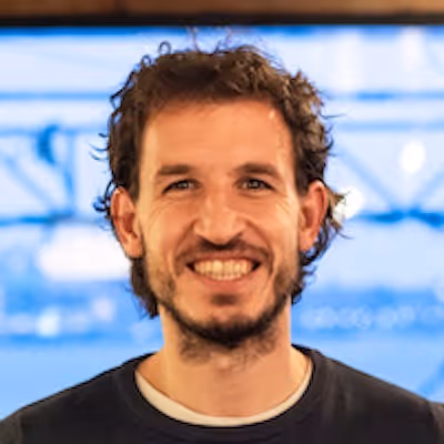Portret van een glimlachende man met krullend haar en een baard, met een wazige achtergrond.