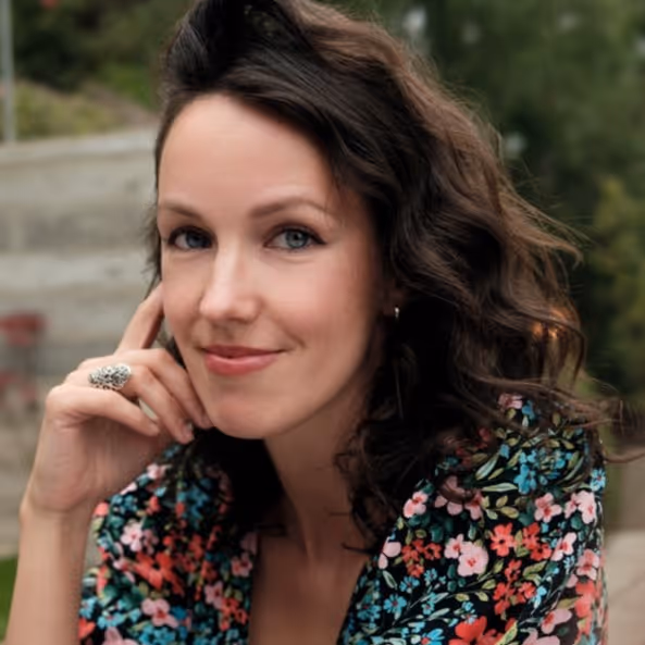 Vrouw met krullend donker haar, glimlachend en licht leunend met hand tegen gezicht, draagt een kleurrijk bloemenpatroon blouse.