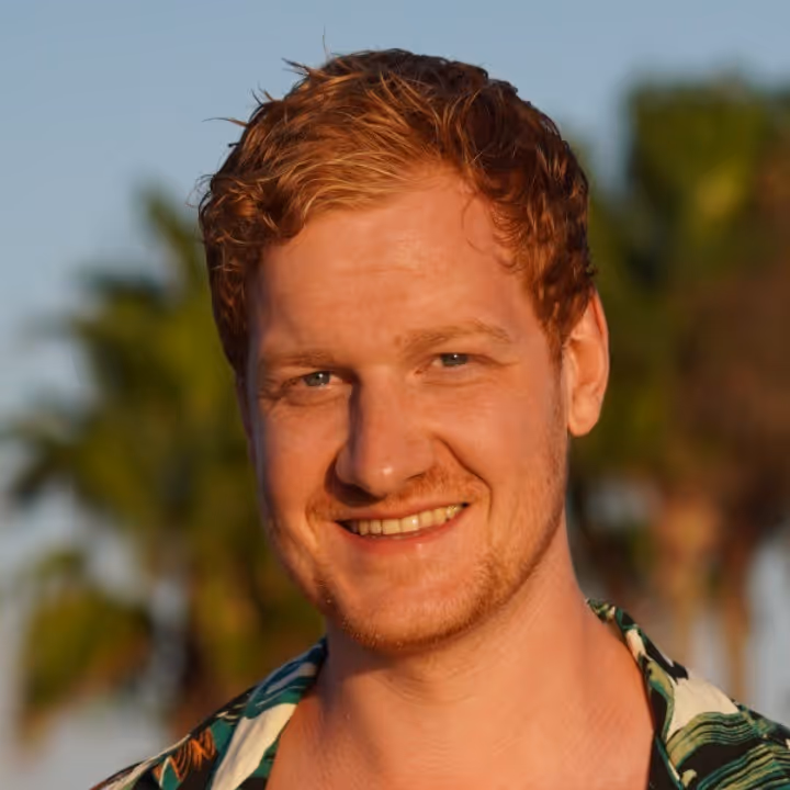 Close-up van een lachende man met rood haar en baard, met palmbomen op de achtergrond.