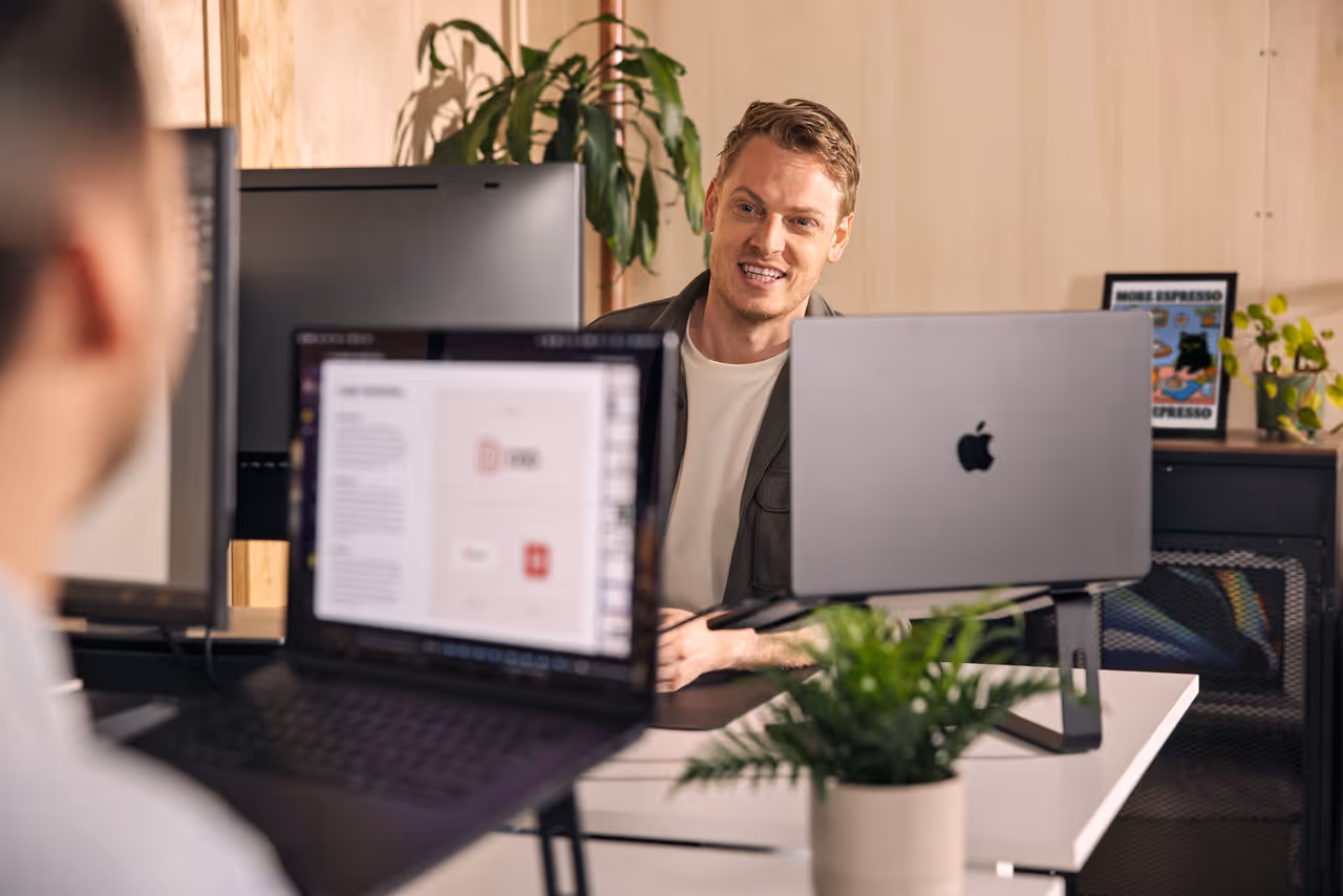 Twee mannen in een moderne kantoorruimte praten achter laptops, met planten en een poster op de achtergrond.