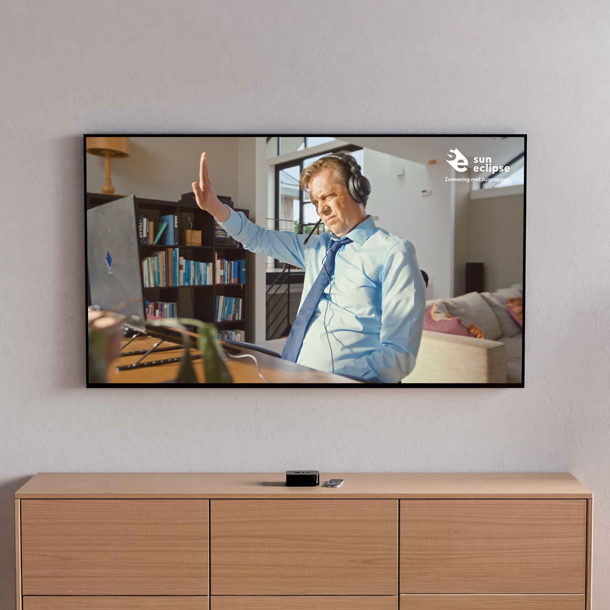 Man met headset doet handgebaar terwijl hij achter een bureau zit met boekenplank en bank op de achtergrond.