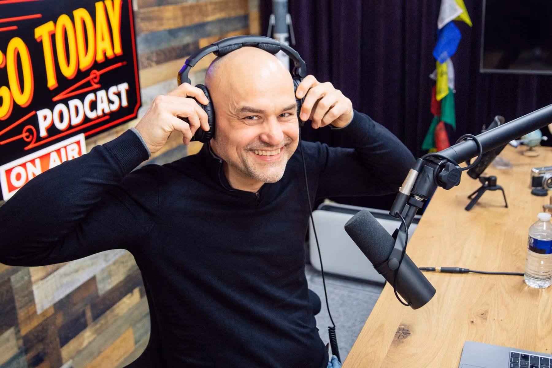 Smiling bald man in a black sweater adjusting headphones in a podcast studio.