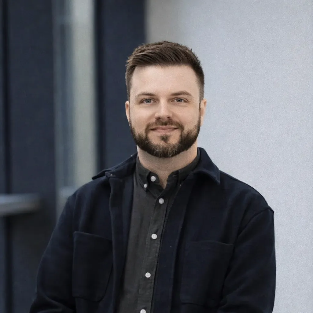 Porträt eines lächelnden Mannes mit kurzem braunem Haar und Bart, der einen dunklen Hemdjacke trägt.