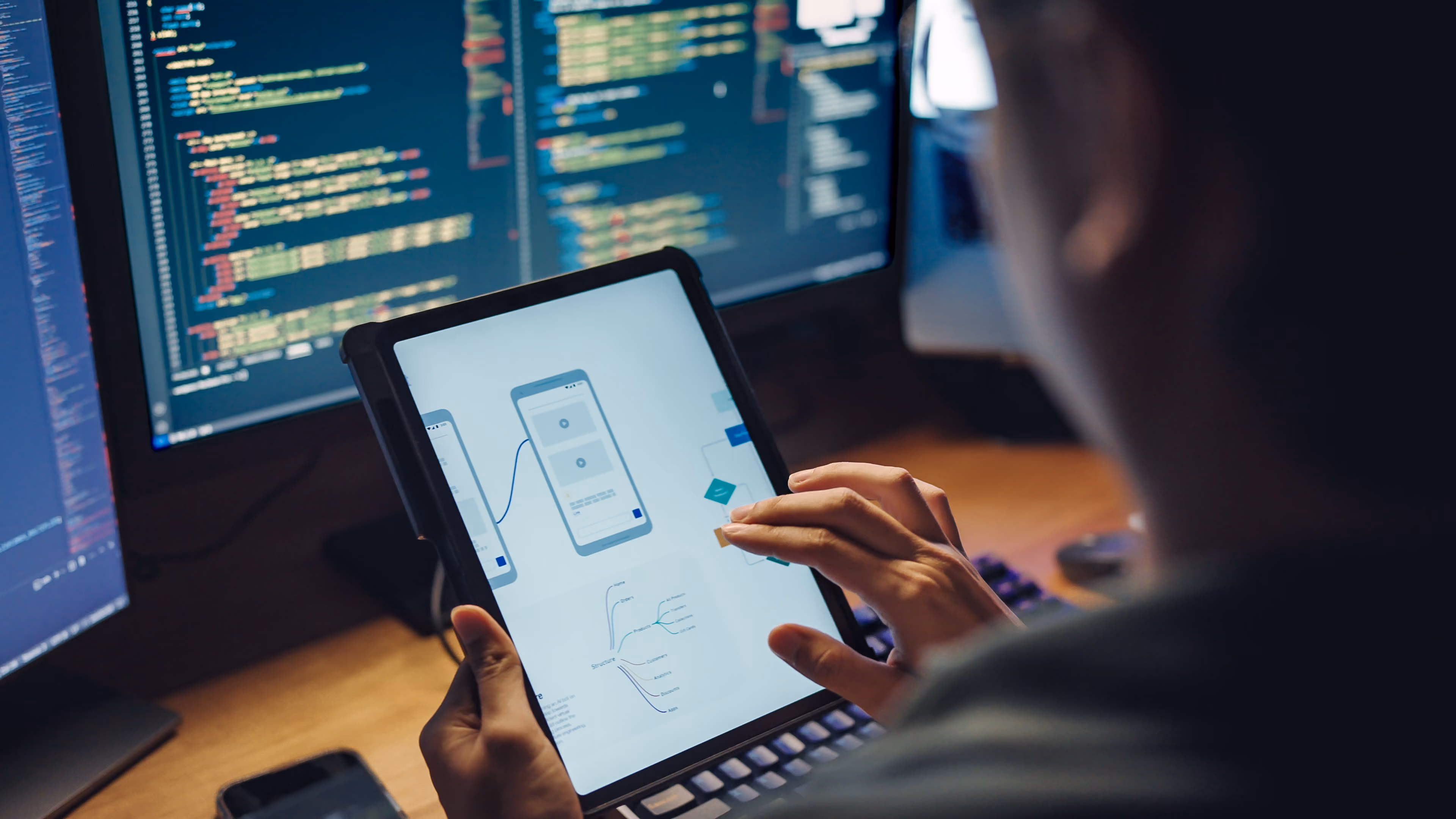 Person interacting with a tablet displaying mobile app design wireframes near computer screens showing code.