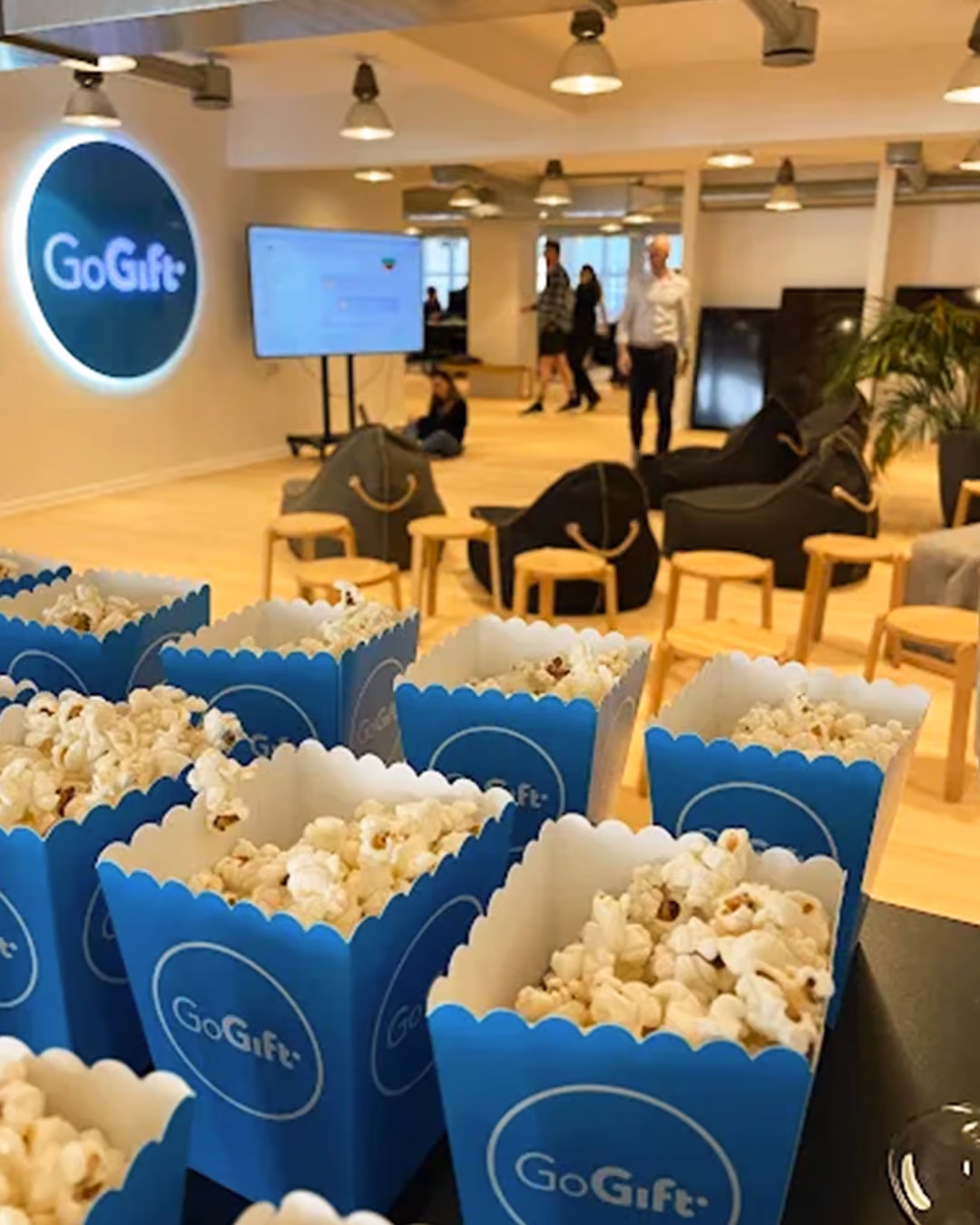 Popcorn in blue GoGift containers in a modern office lounge with people and bean bag chairs.