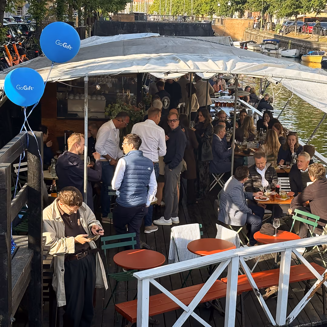Crowd socializing and dining under a canopy on a boat docked by a canal.
