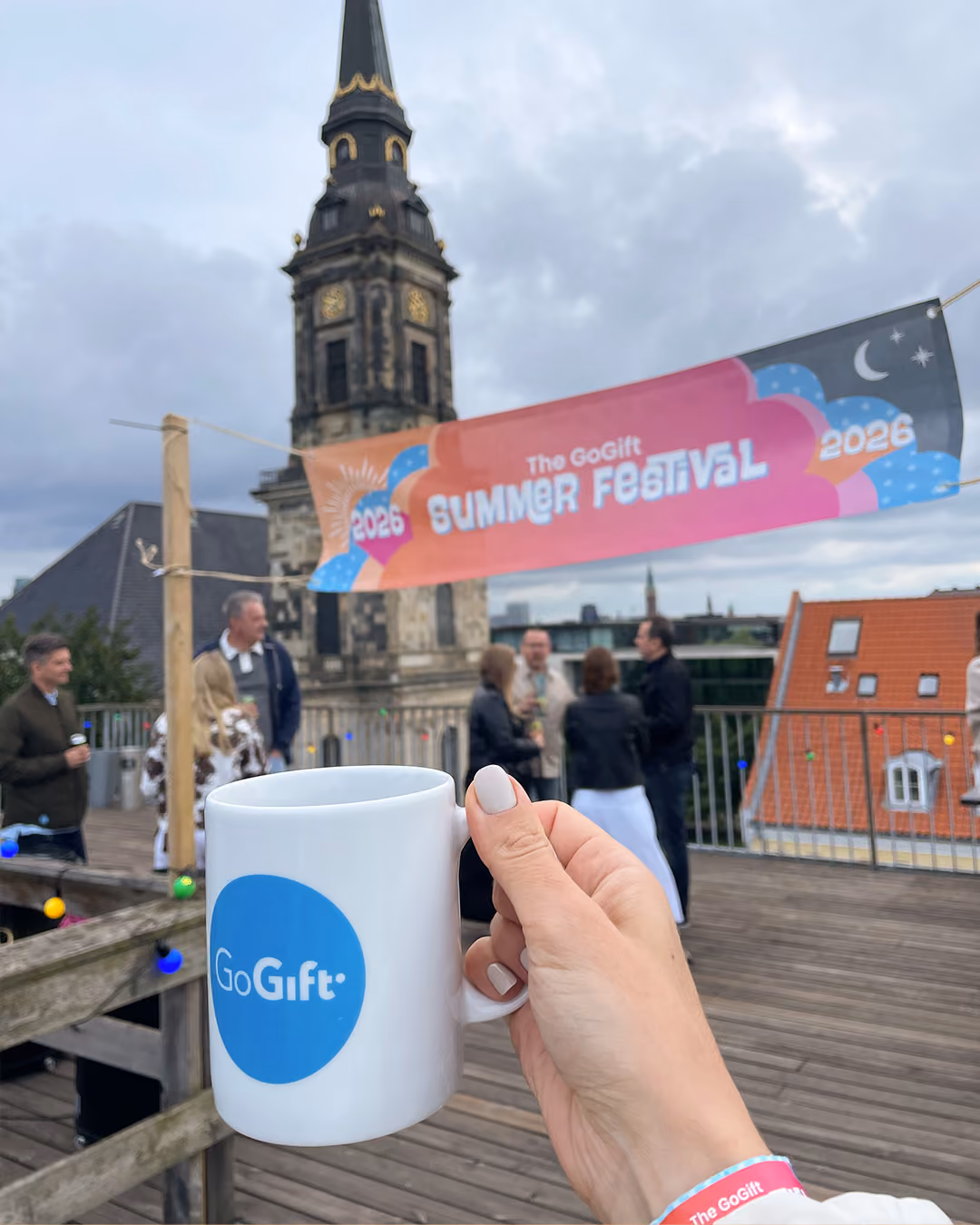 Hand holding a GoGift mug at an outdoor summer festival with a tall clock tower and crowd.