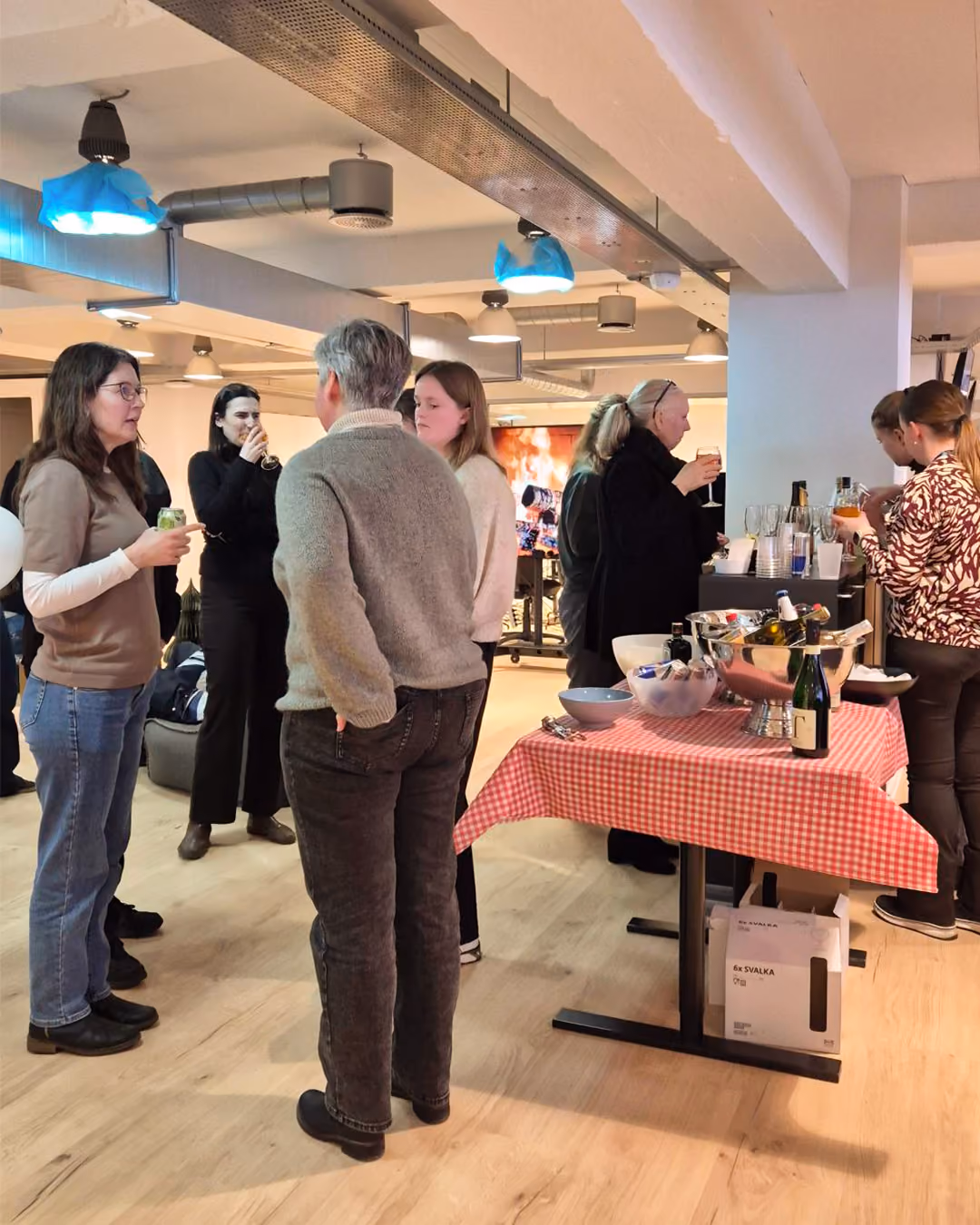 People socializing and drinking near a table with drinks and snacks on a red checkered tablecloth.