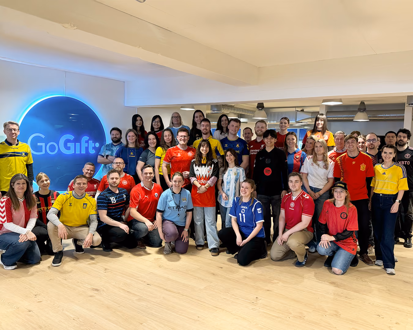 A large group of people posing indoors wearing various soccer jerseys in front of a GoGift sign.