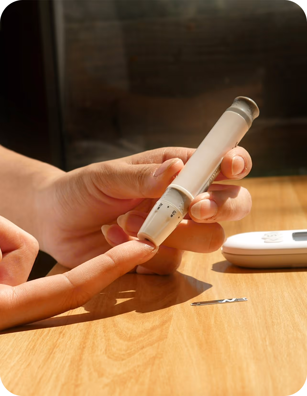 Person using a lancet device to prick their finger for a blood glucose test with a glucose meter and lancet on a wooden table.