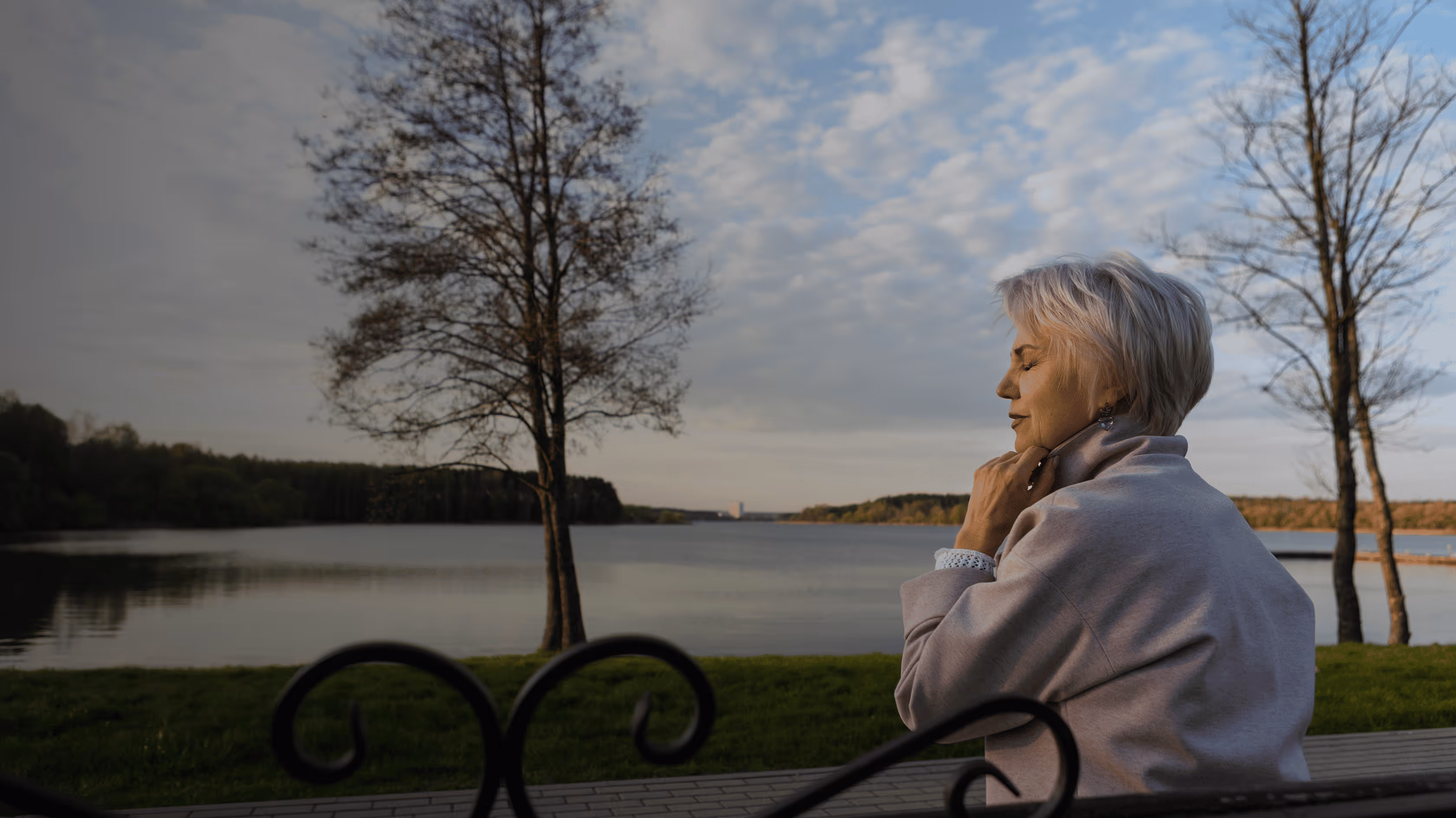 Smiling older woman with short white hair stretching her arms behind her head outdoors in a green park.