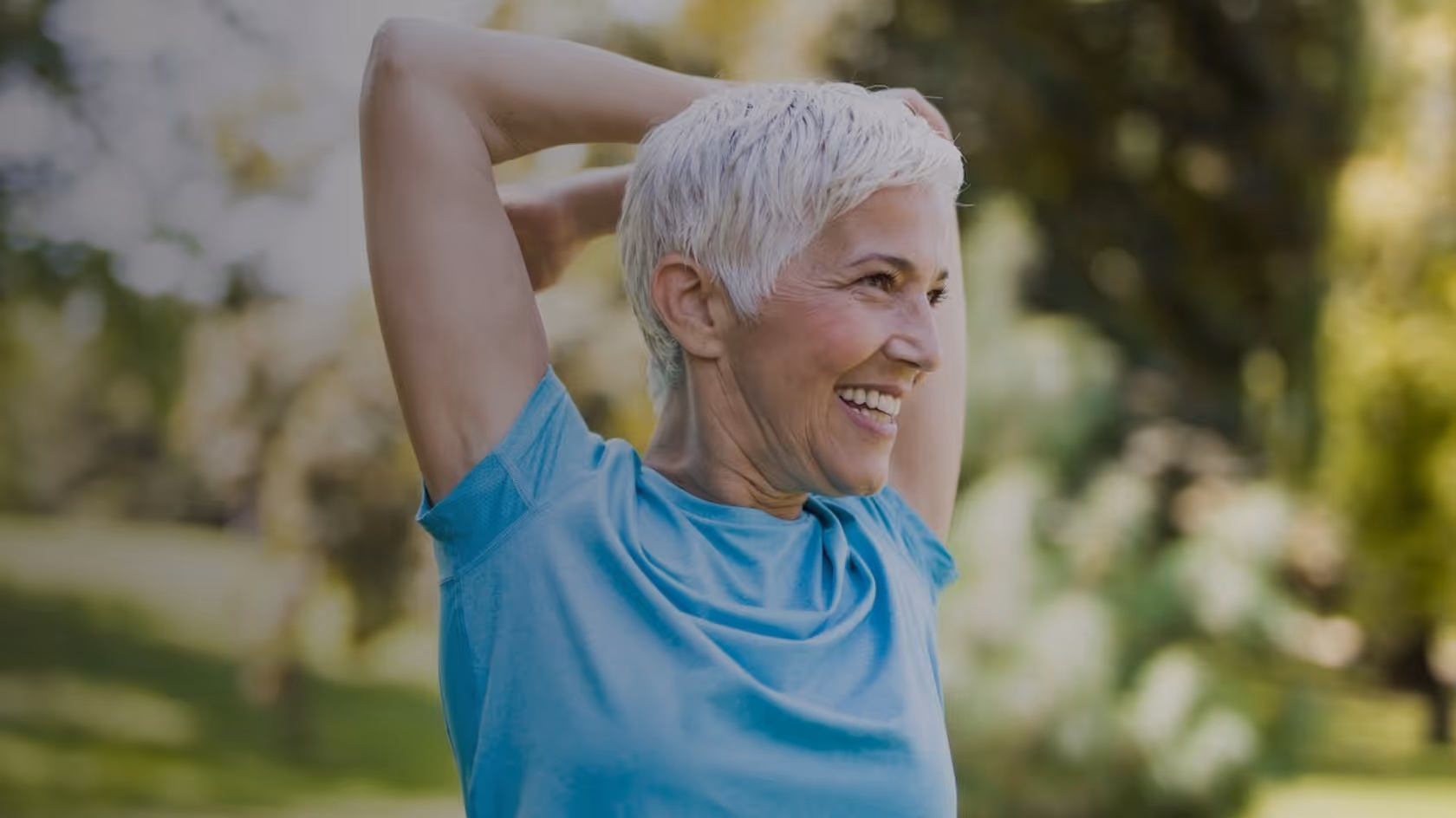 Smiling older woman stretching outdoors in a blue shirt with greenery in the background.