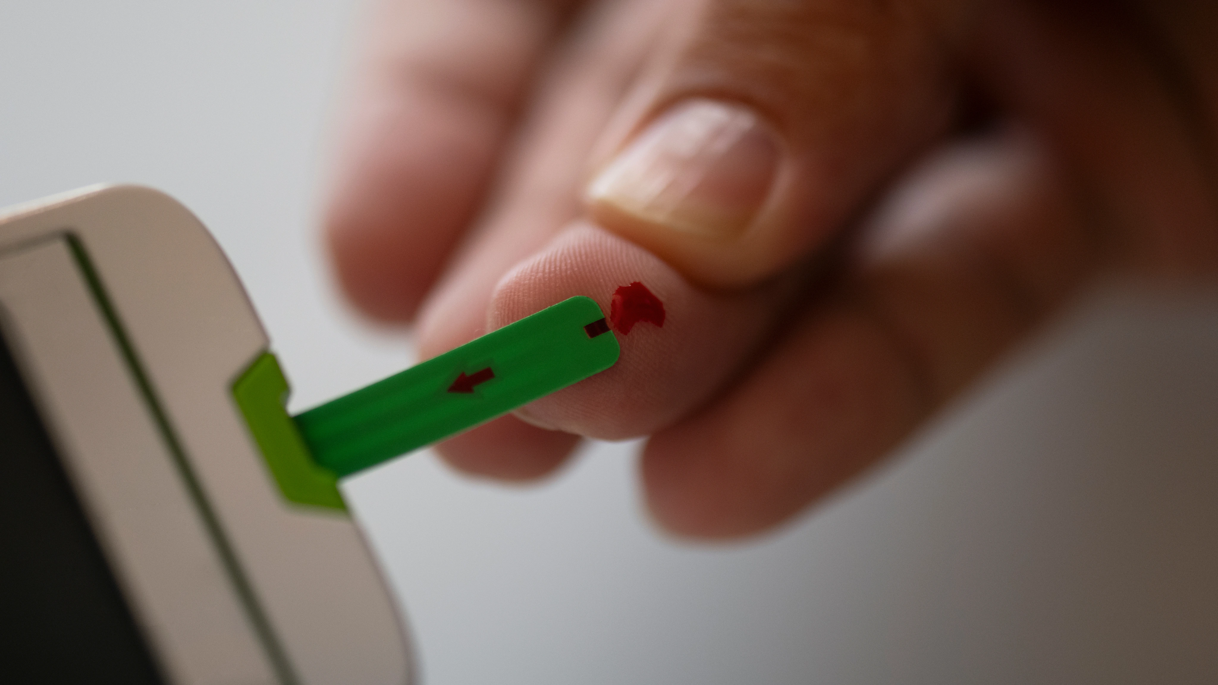 Finger with a drop of blood being tested using a green test strip inserted into a glucose meter.