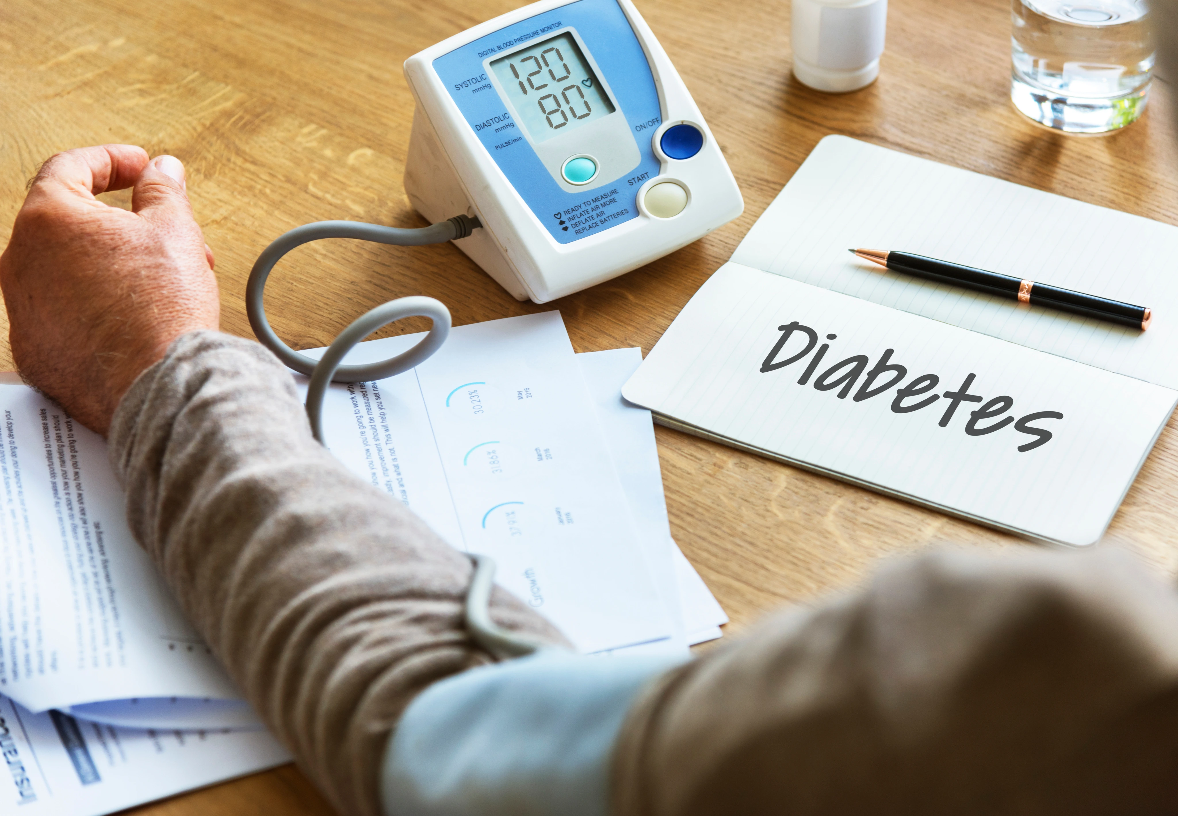 Person measuring blood pressure with digital monitor showing 120/80 next to notebook with 'Diabetes' written on it.