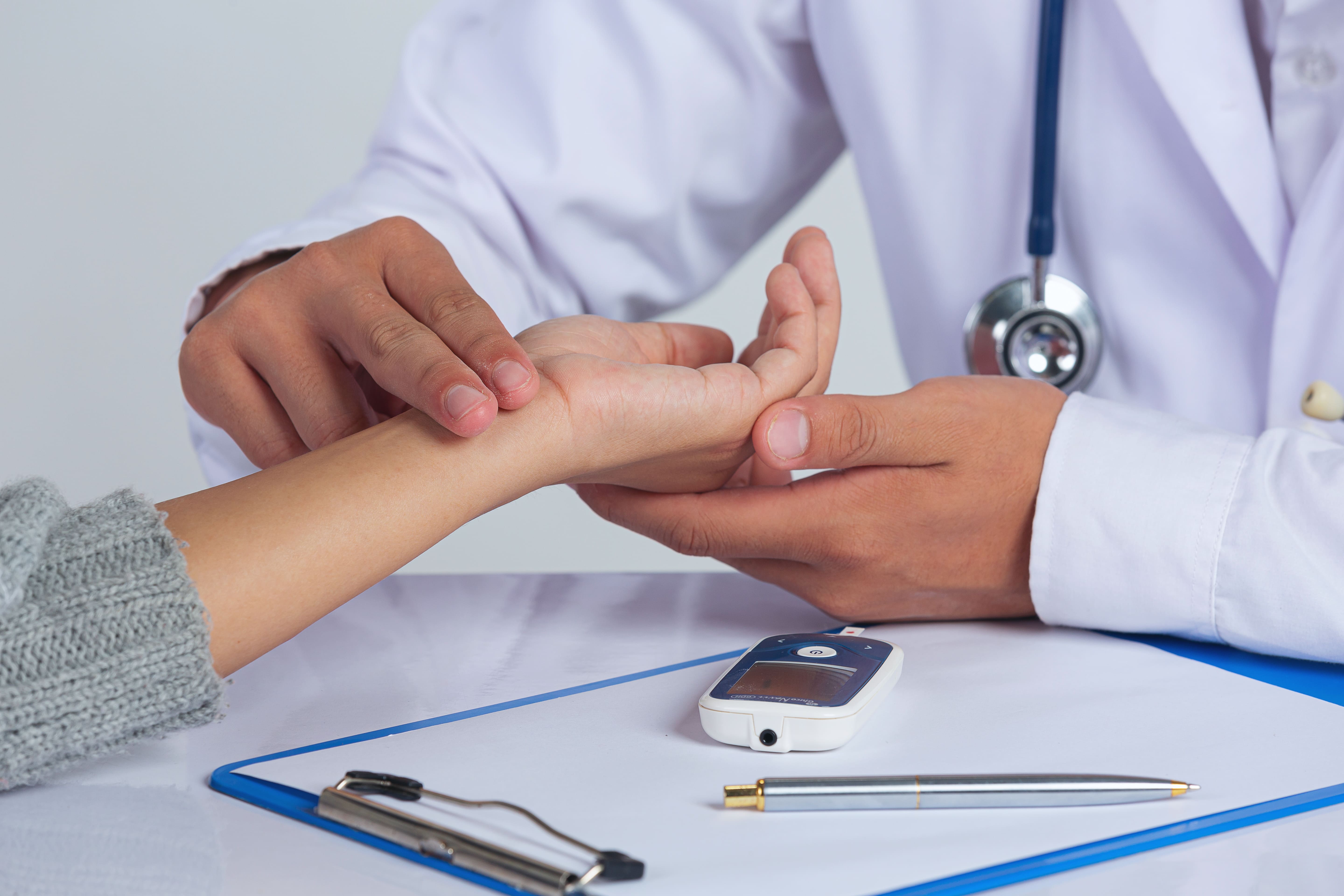 Doctor checking patient's wrist pulse with glucometer and pen on clipboard on table.