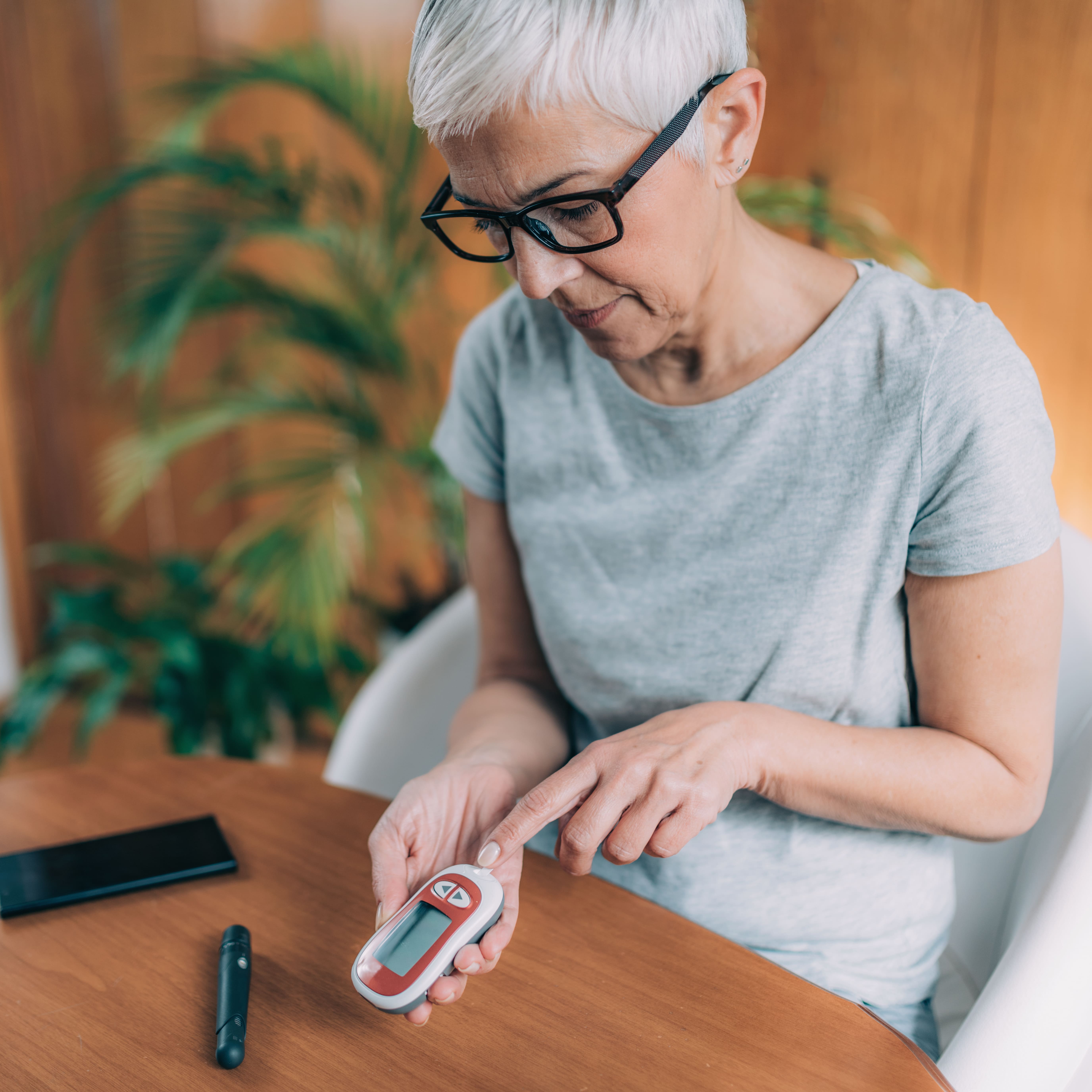 Senior woman with glasses using a blood glucose meter to check her blood sugar at a wooden table.